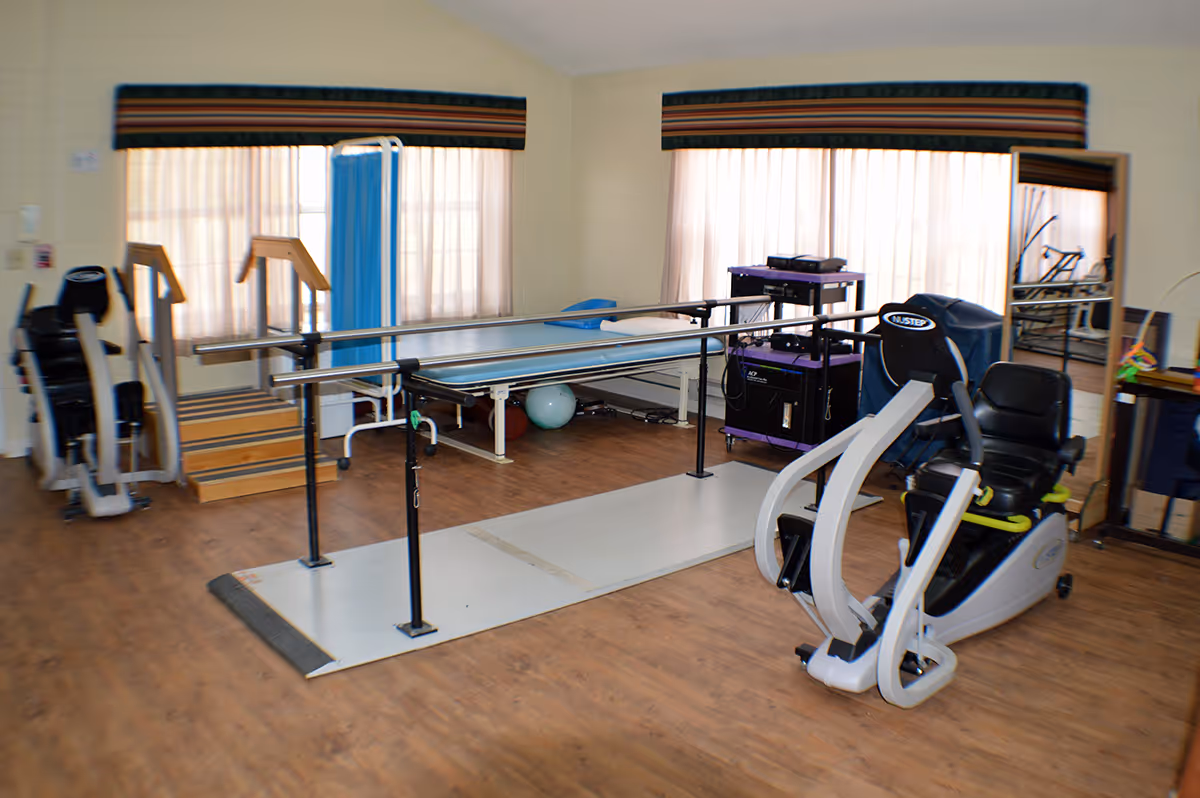 A rehabilitation room with exercise equipment including parallel bars for walking practice, a recumbent exercise bike, a treatment table, and a small set of stairs. The room has large windows with curtains, wooden flooring, and a mirror on one wall.