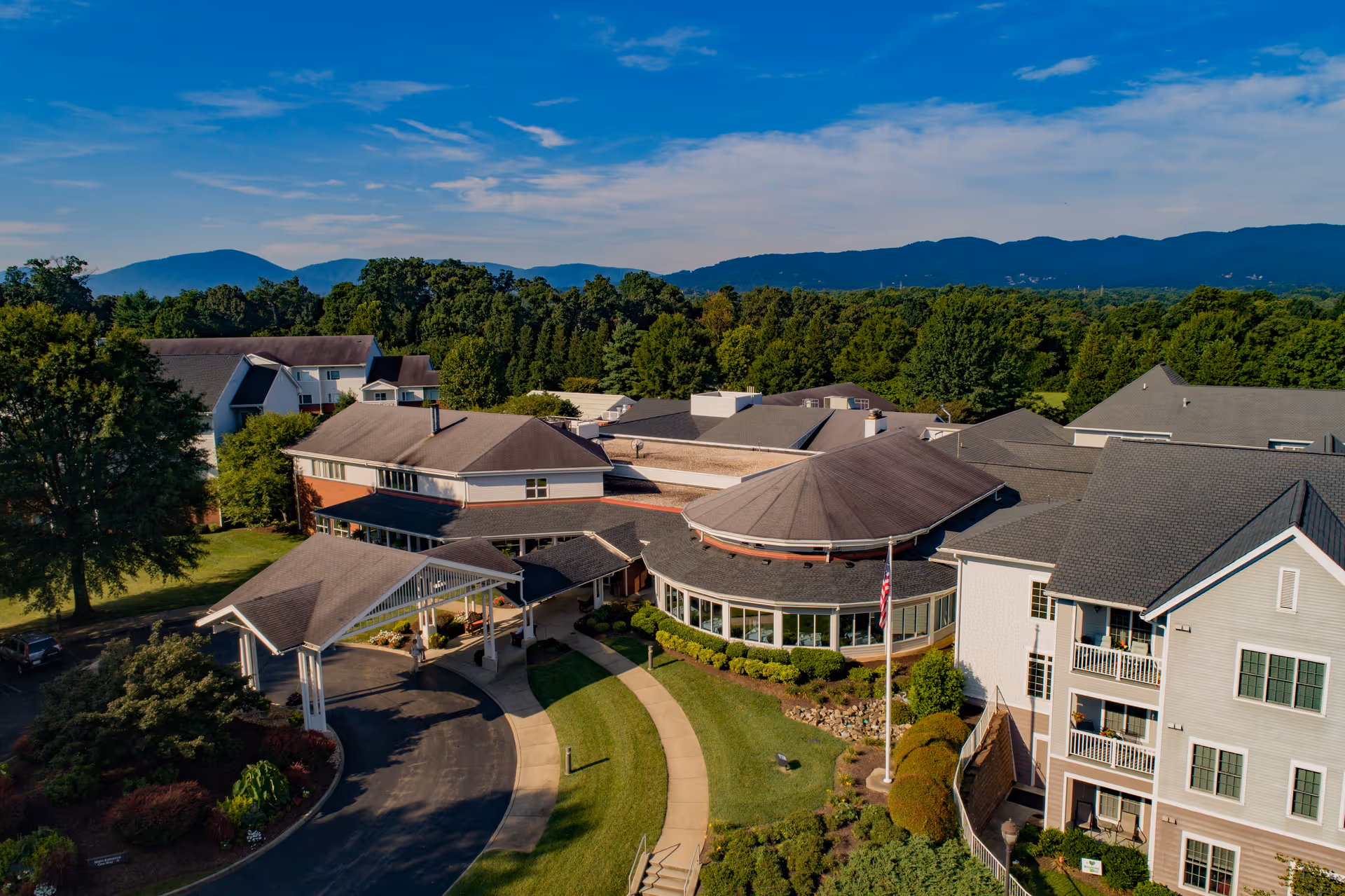 Aerial view of Brandon Oaks Life Plan Community showing multiple connected buildings with pitched roofs, surrounded by green trees and landscaped gardens, with mountains visible in the background under a partly cloudy blue sky.