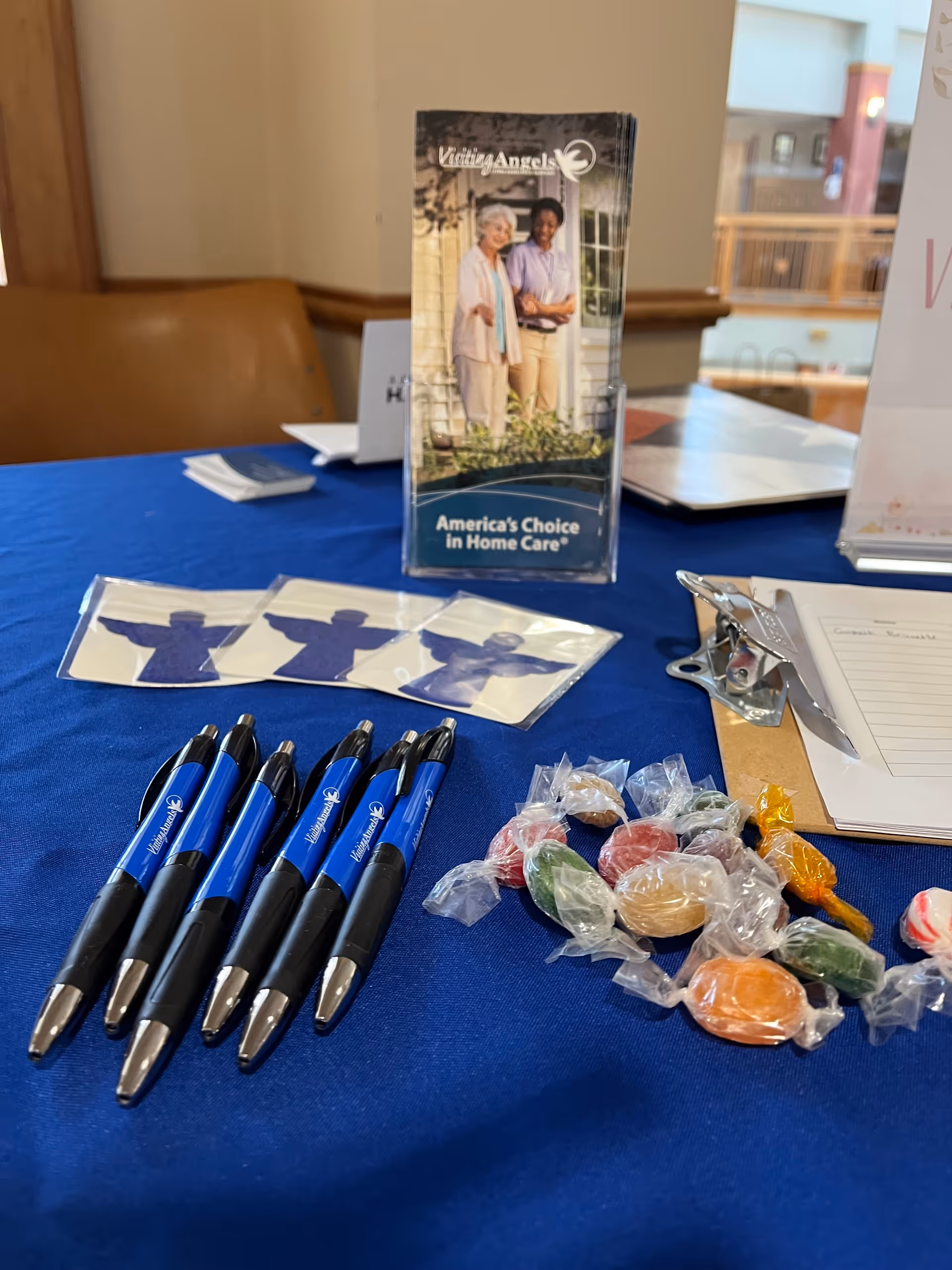 Promotional table with pens, candies, and a Visiting Angels brochure in an indoor lobby.