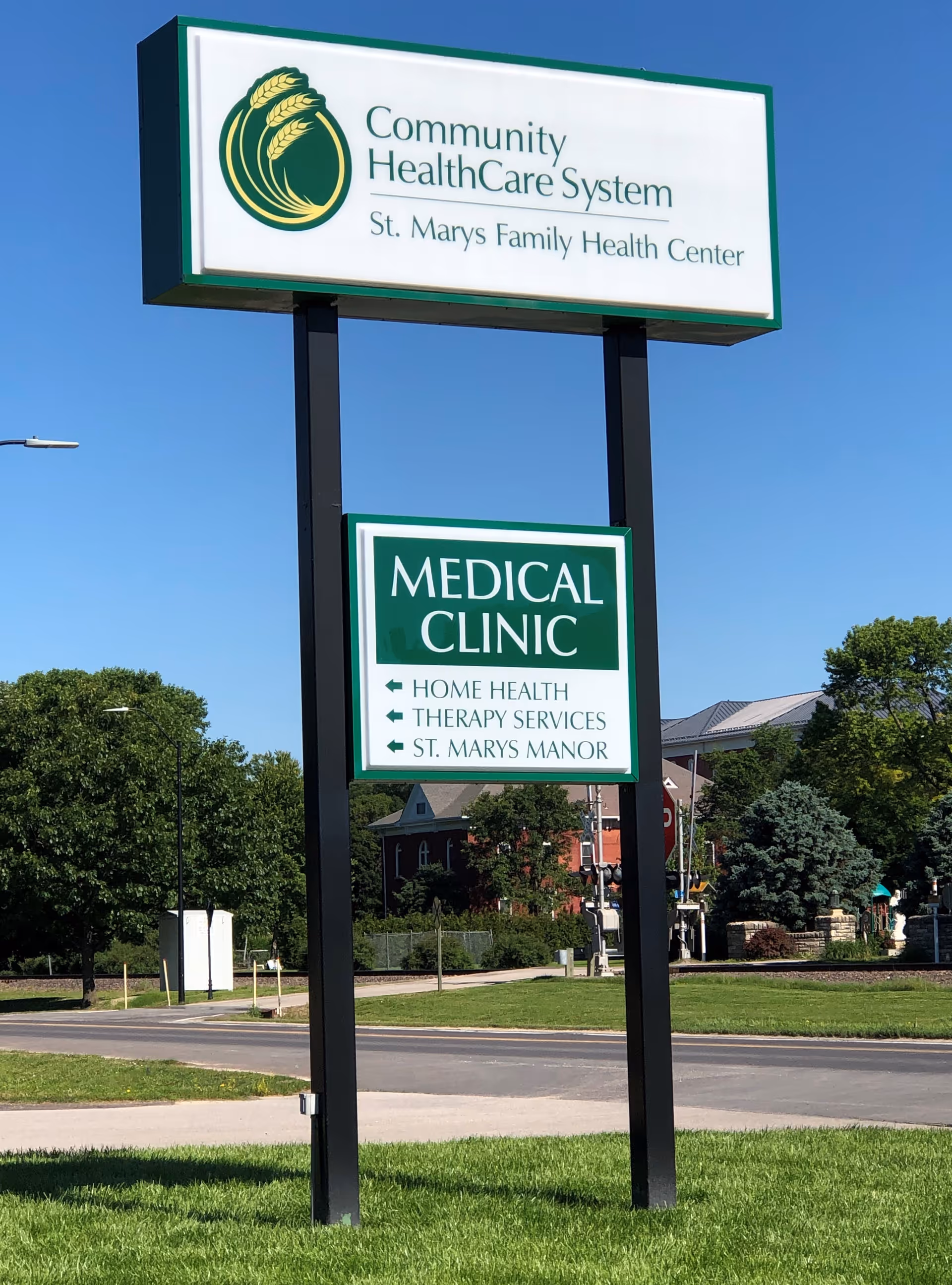 Outdoor view of a tall sign for Community HealthCare System St. Marys Family Health Center with a smaller sign below indicating Medical Clinic, Home Health, Therapy Services, and St. Marys Manor. The sign is set on a grassy area near a road with trees and buildings in the background under a clear blue sky.