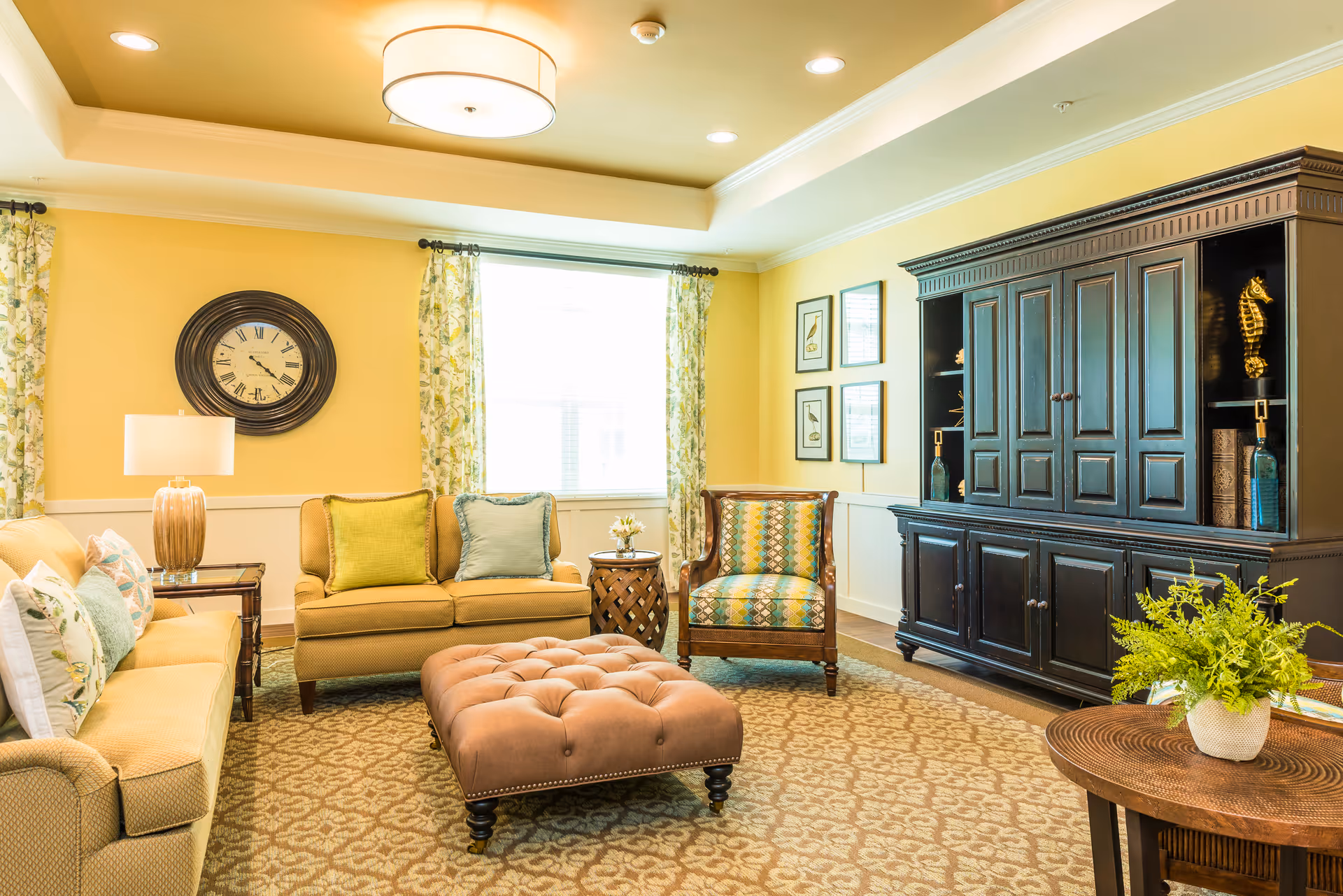 Cozy senior living lounge with beige sofas, patterned chairs, tufted ottoman, and dark cabinet.