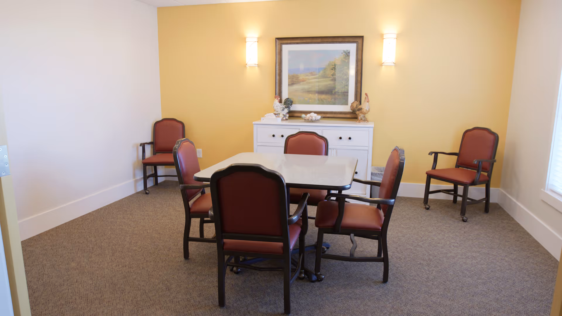 Small dining room with a square table surrounded by red-upholstered chairs, a white sideboard and framed artwork on a yellow accent wall.