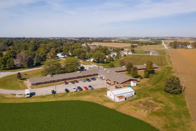 Aerial view of Falls City Nursing and Rehabilitation Center, a single-story building with a parking lot containing several cars, surrounded by green fields, trees, and rural landscape under a partly cloudy sky.