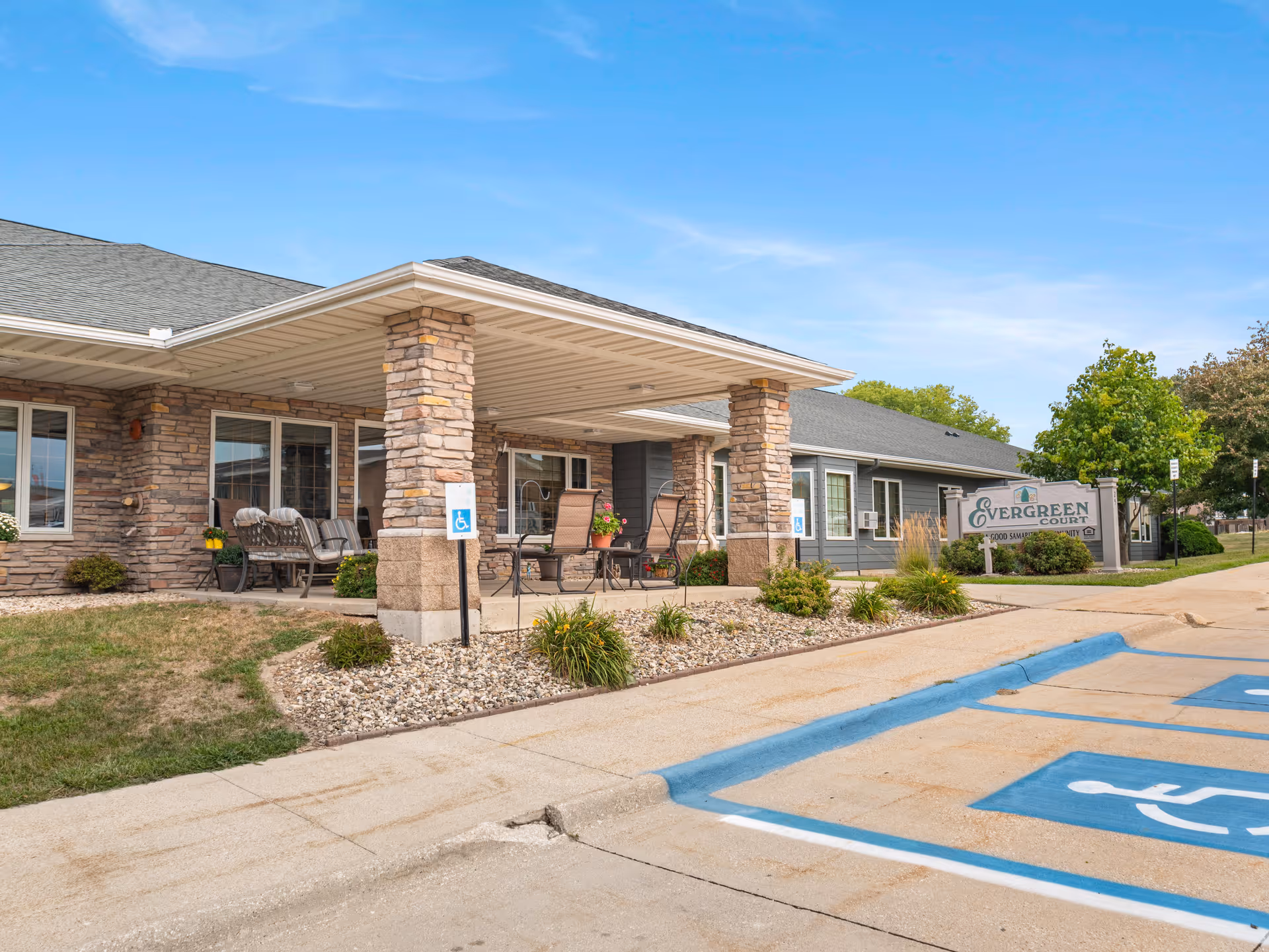 Exterior view of Good Samaritan Society - Waukon showing a covered patio area with stone pillars, outdoor seating, and a sign that reads Evergreen Court. There are handicap parking spaces in the foreground and some landscaping with bushes and grass around the building.