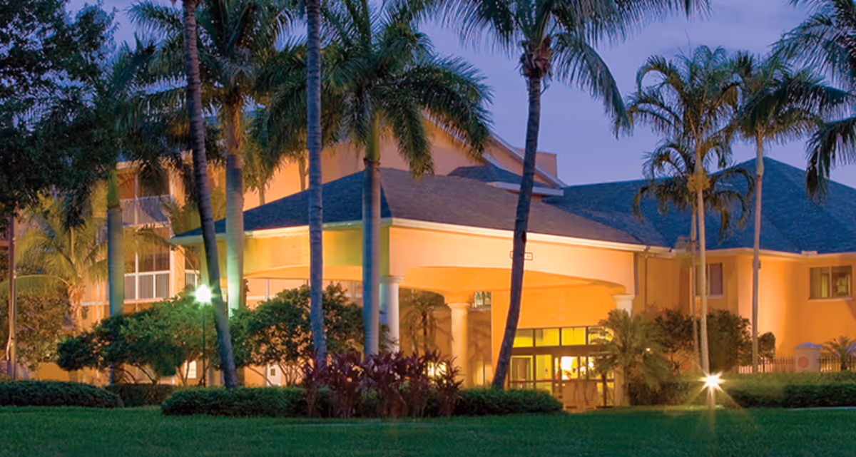 Exterior view of a senior living facility building at dusk with palm trees and well-maintained landscaping in front. The building is illuminated with warm lights and has a covered entrance supported by columns.