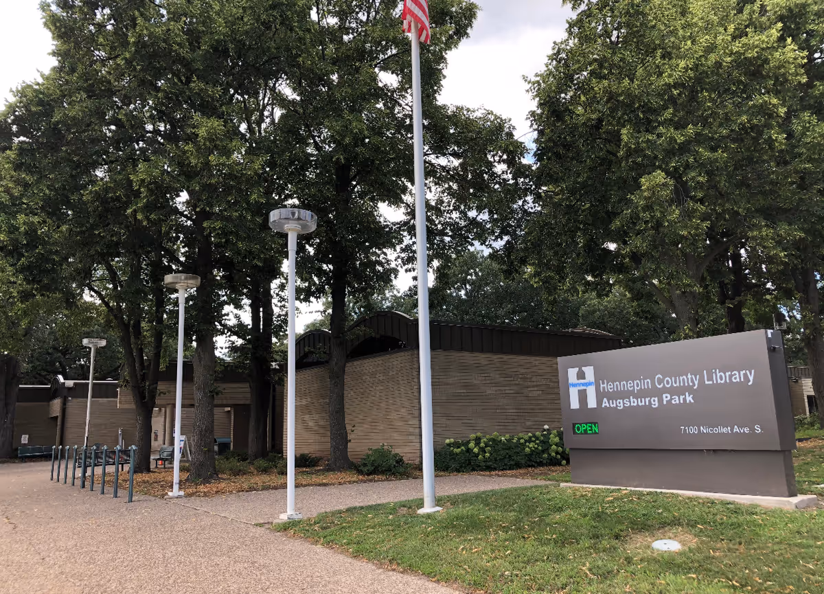 Exterior view of the Hennepin County Library Augsburg Park building surrounded by trees and a pathway with lamp posts and bike racks. A sign in front displays the library name, address, and an open status.