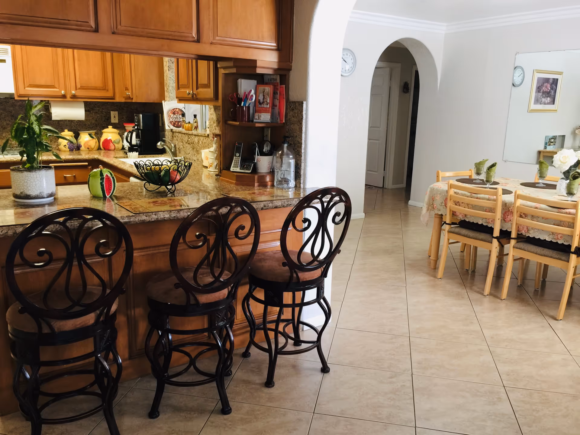 Interior view of a kitchen and dining area in a senior living facility. The kitchen features wooden cabinets, a granite countertop with three ornate black metal bar stools, a coffee maker, and decorative items including a plant and fruit bowl. The dining area has a table covered with a floral tablecloth, six wooden chairs with black cushions, green napkins, and a vase with white flowers. The floor is tiled, and there is an arched doorway leading to another room.