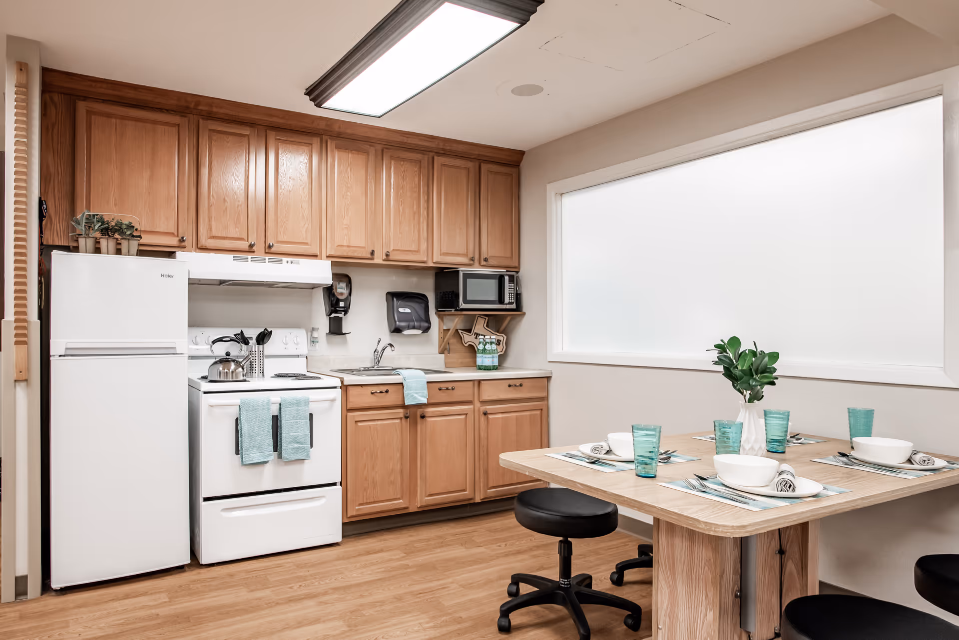A clean and well-lit kitchen area with wooden cabinets, a white refrigerator, stove, microwave, and sink. A small dining table is set with four place settings including bowls, glasses, and napkins, with a small green plant centerpiece. The floor is wooden and there is a large frosted window on the wall.