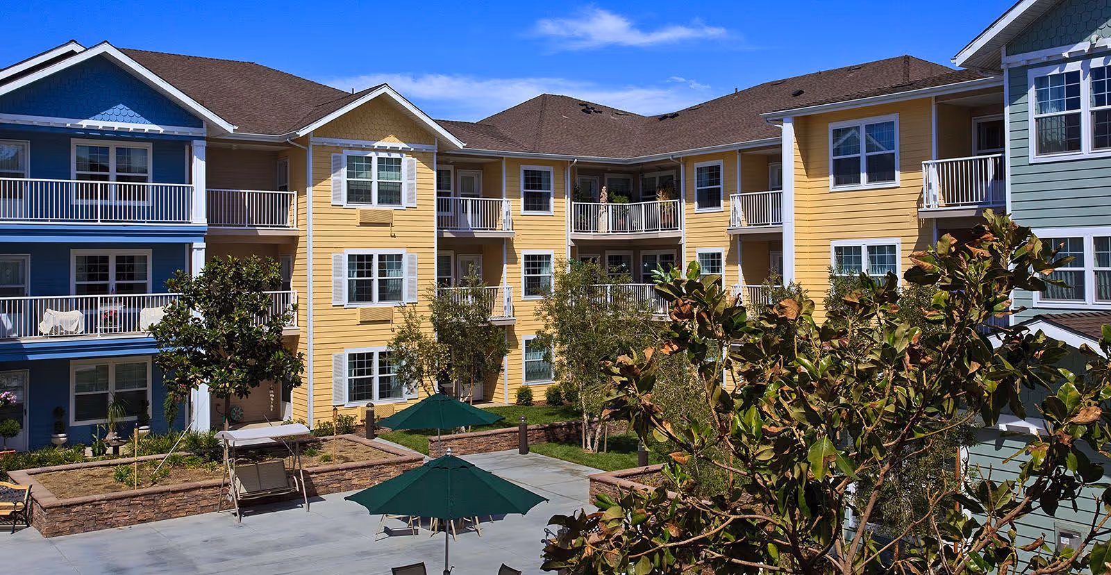 Three-story pastel-colored apartment buildings surrounding a landscaped courtyard with patio umbrellas and balconies.