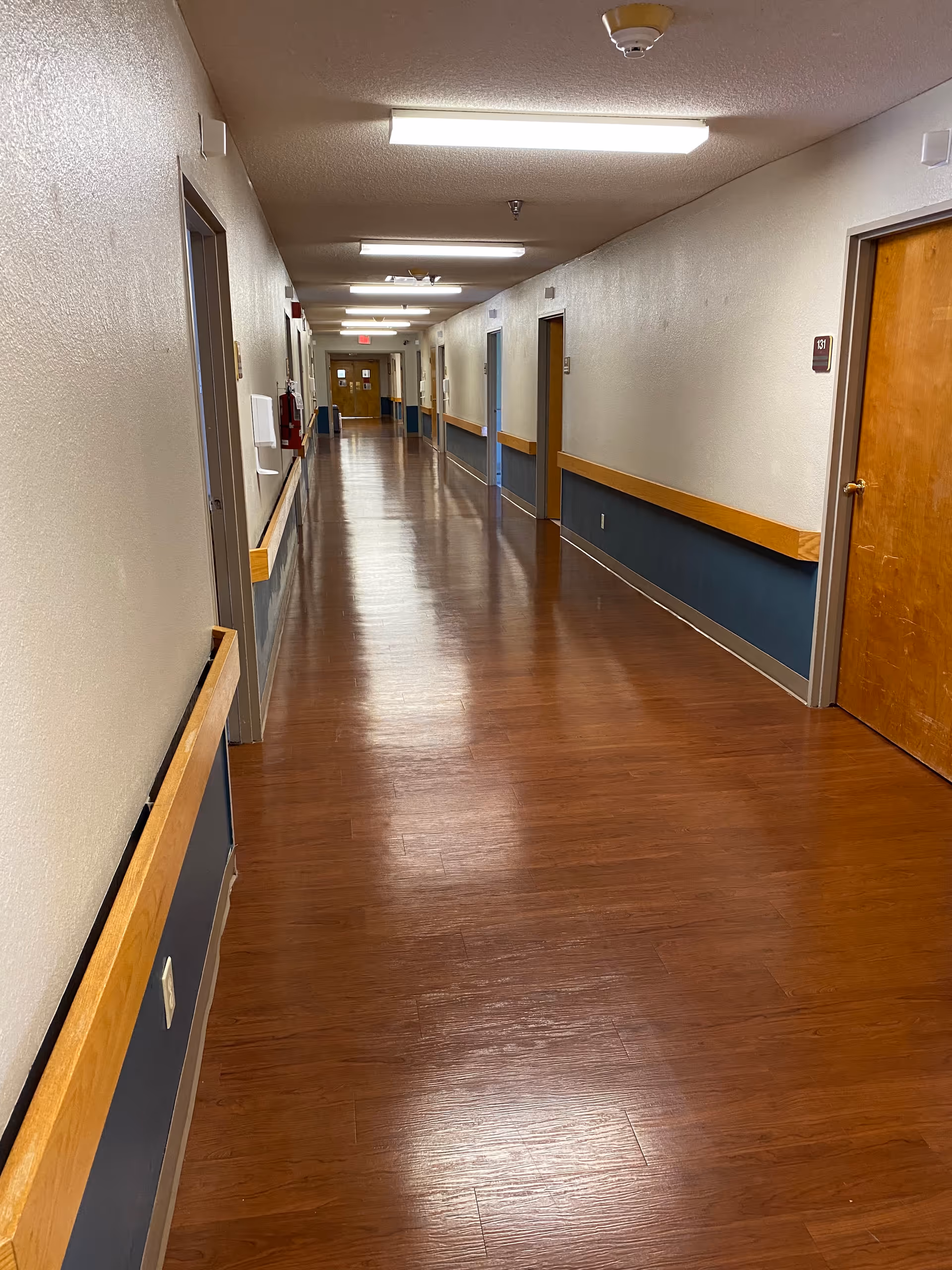 Long, well-lit hallway in a senior living facility, with doors along both sides and wooden flooring.