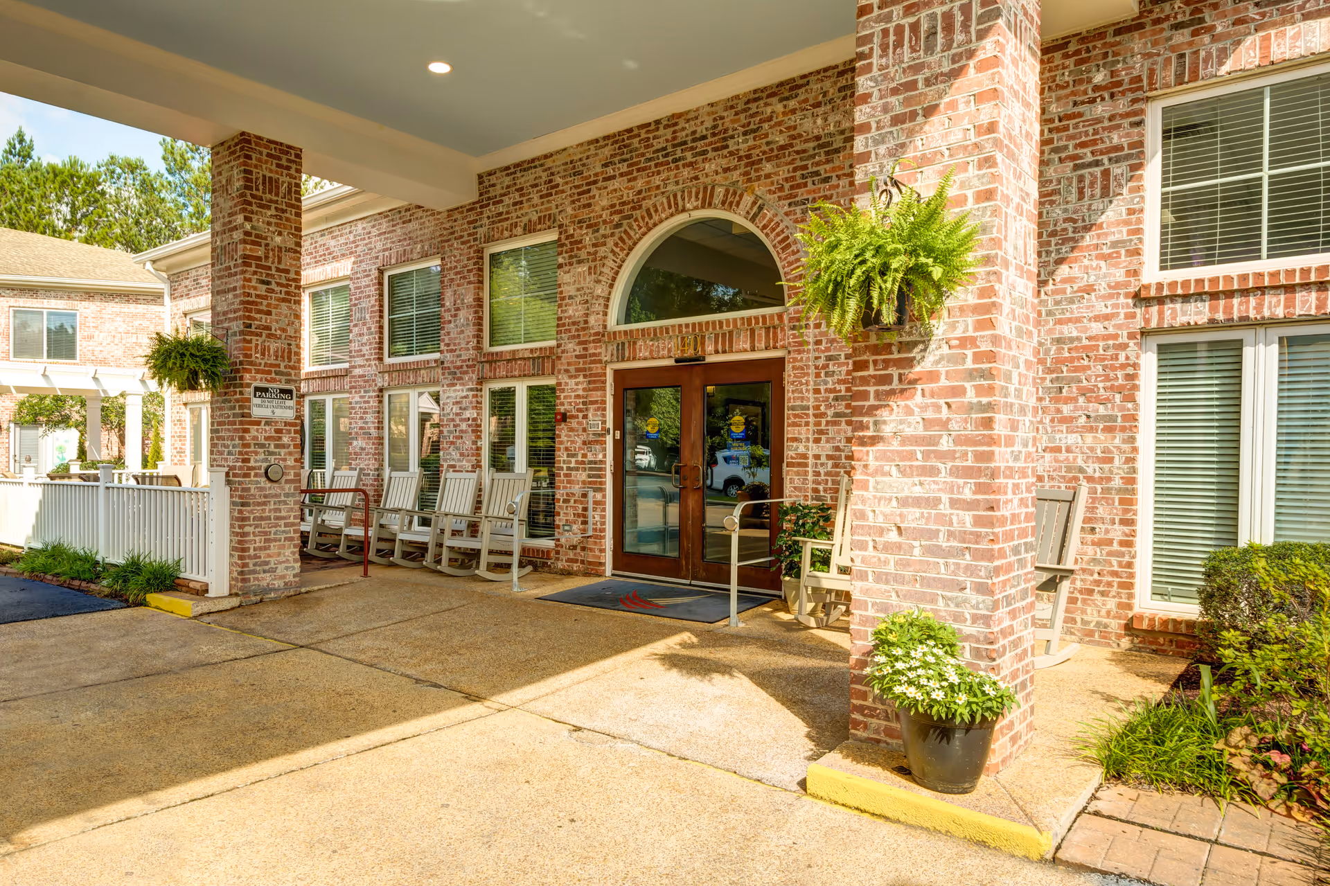 Entrance of Castlewoods Place featuring a covered porch with brick pillars, several rocking chairs, hanging plants, potted flowers, and double glass doors with an arched window above.