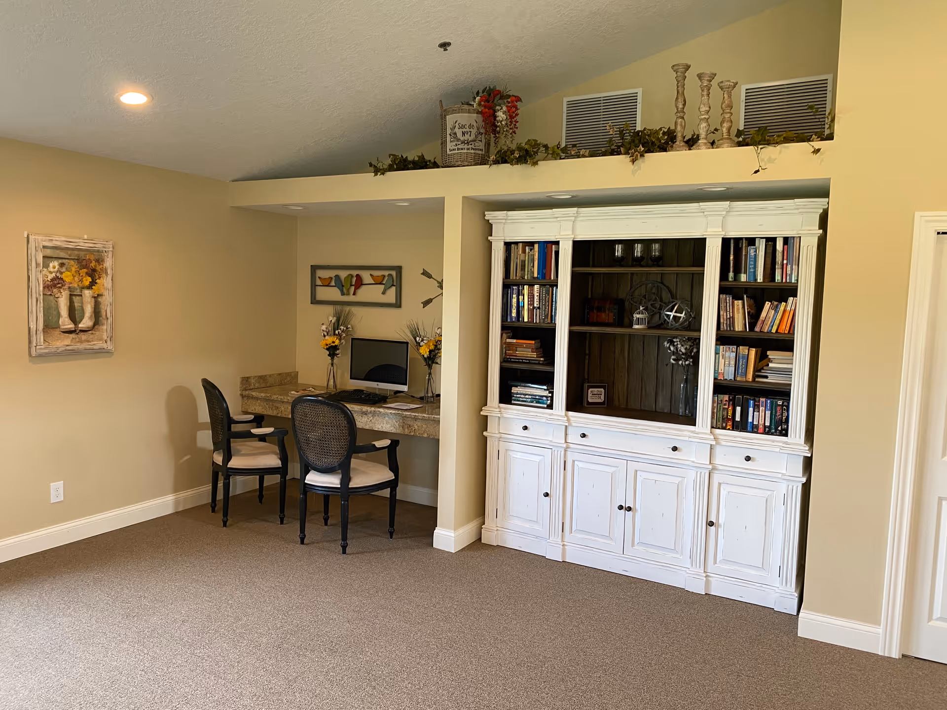 A cozy interior corner of a senior living community featuring a built-in desk with two chairs and a computer, a large white wooden bookshelf filled with books and decorative items, beige walls, and carpeted floor. Above the bookshelf, there are decorative plants and candlesticks.