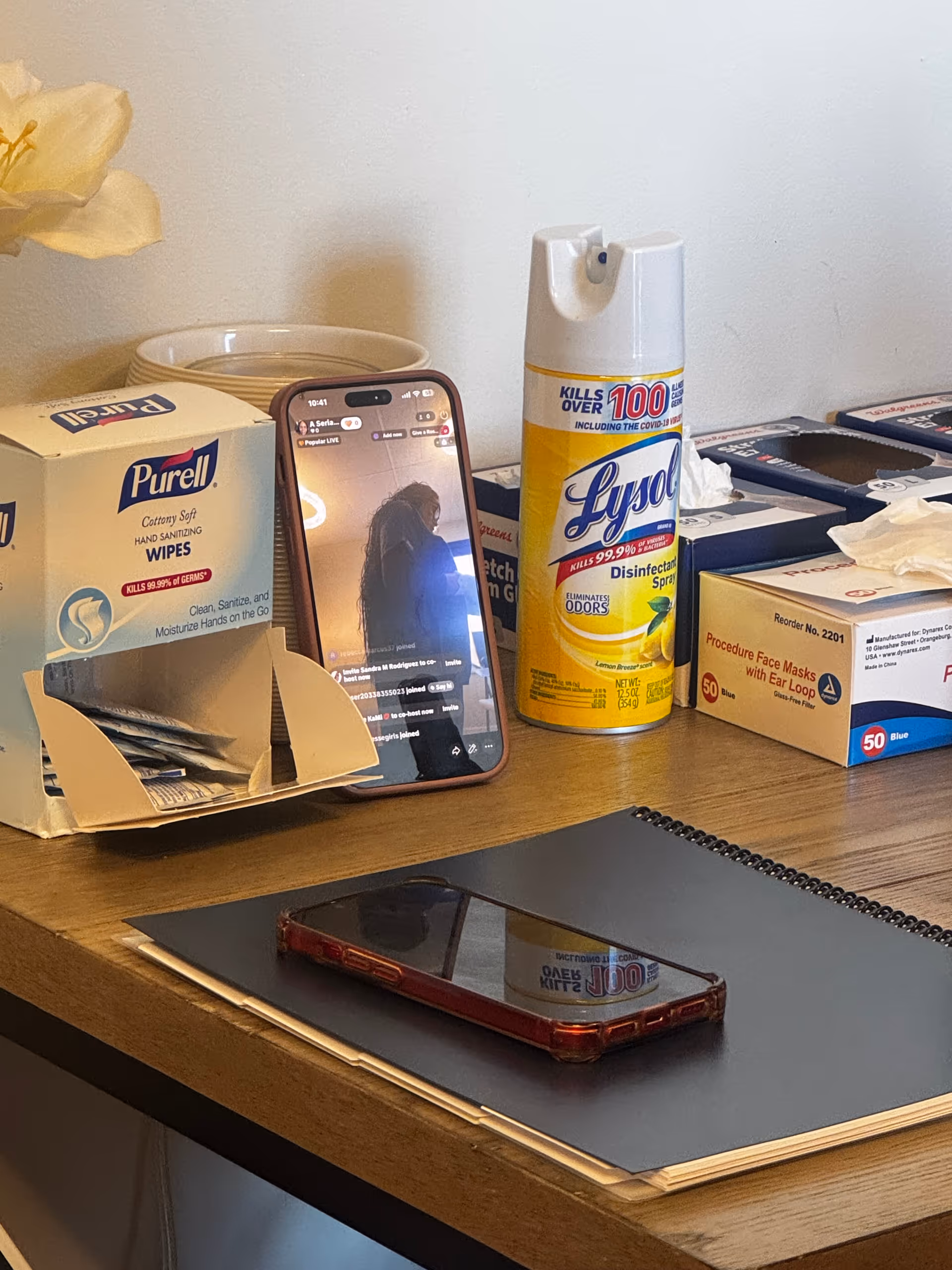 Office desk cluttered with wipes, disinfectant spray, tissue box, notebooks, and a smartphone.
