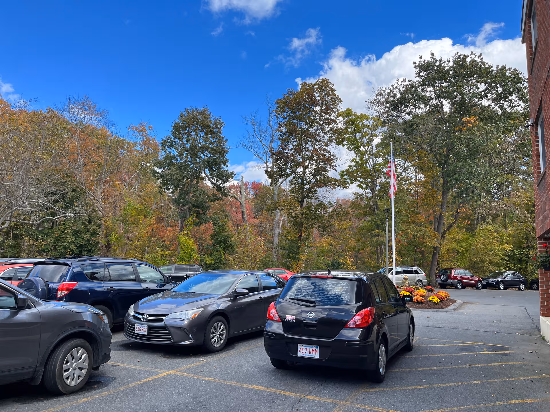 Parking lot with cars, autumn trees, flagpole, and brick building edge on the right.