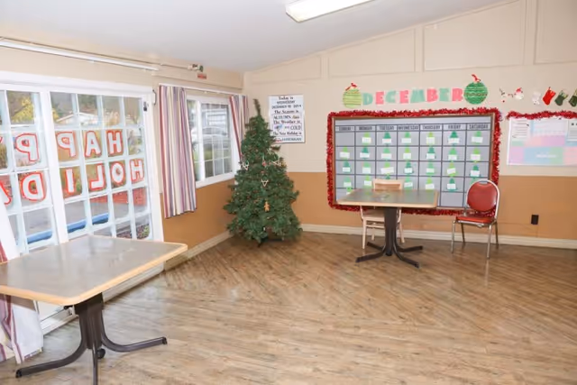 A communal activity room with two tables and chairs, a small Christmas tree, sliding glass doors decorated with "Happy Holidays", and a December bulletin board on the wall.