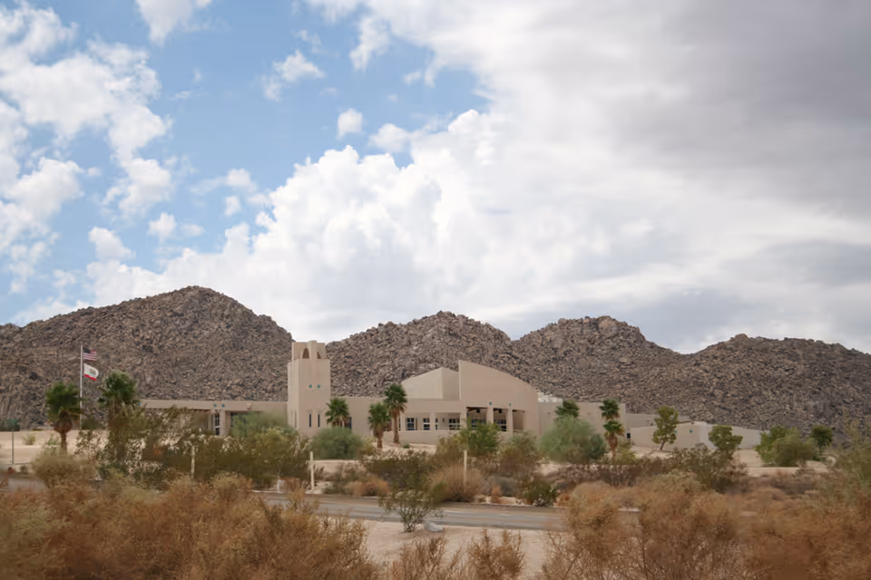 Low-slung beige care facility with palm trees in front set against rocky desert hills under a partly cloudy sky.