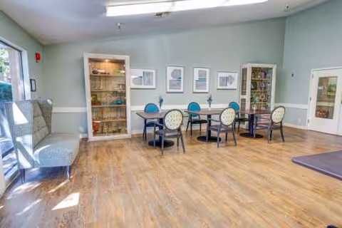 Empty senior living dining area with tables, chairs, a cabinet, and pale walls.
