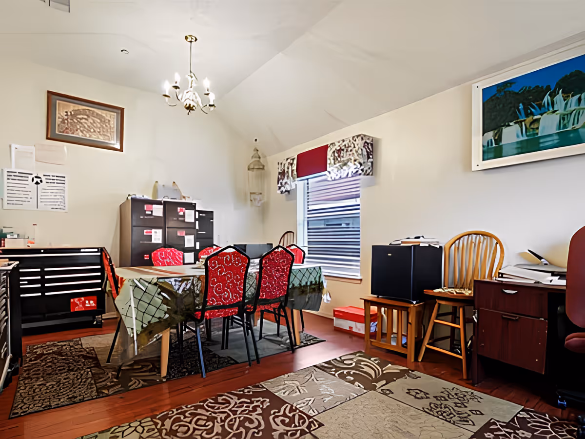 A bright interior dining area with a table surrounded by red-upholstered chairs, filing cabinets, and office furniture.