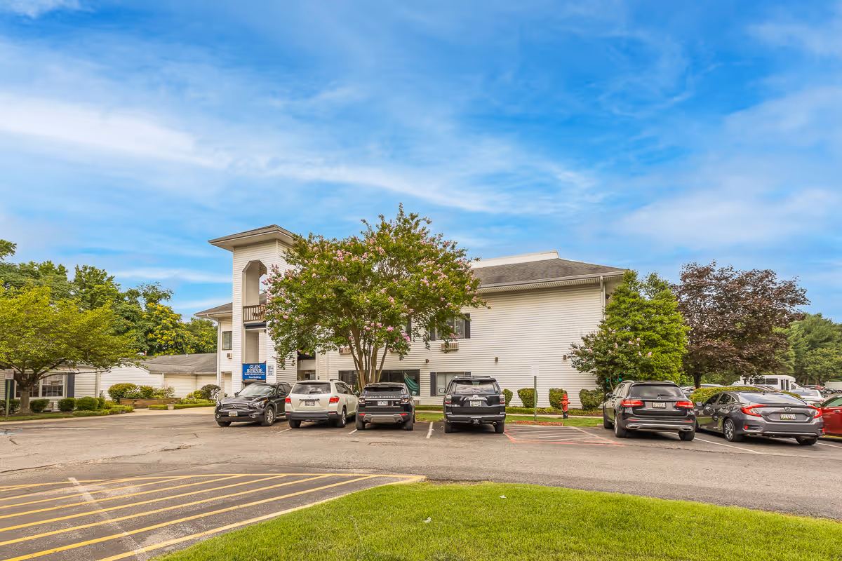 Exterior view of a two-story white building with a parking lot in front. Several cars are parked, and there are trees and green landscaping around the building under a partly cloudy blue sky.