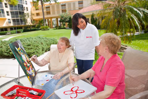 Caregiver assists two elderly residents painting outdoors in a sunny courtyard.