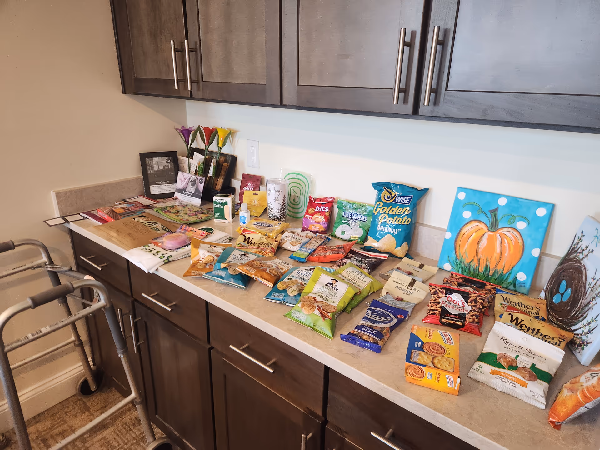 Counter filled with assorted snack bags beside a walker and dark cabinets.