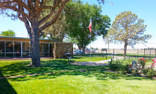 A well-maintained outdoor garden area at Sunset Villa Care Center featuring green grass, trees, a flagpole with an American flag, a stone building with large windows, and a fenced area with flowers and shrubs.
