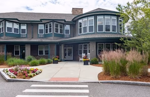 Front exterior view of a two-story senior living facility building with large windows, a covered entrance, landscaped flower beds, and ornamental grasses under a partly cloudy sky.