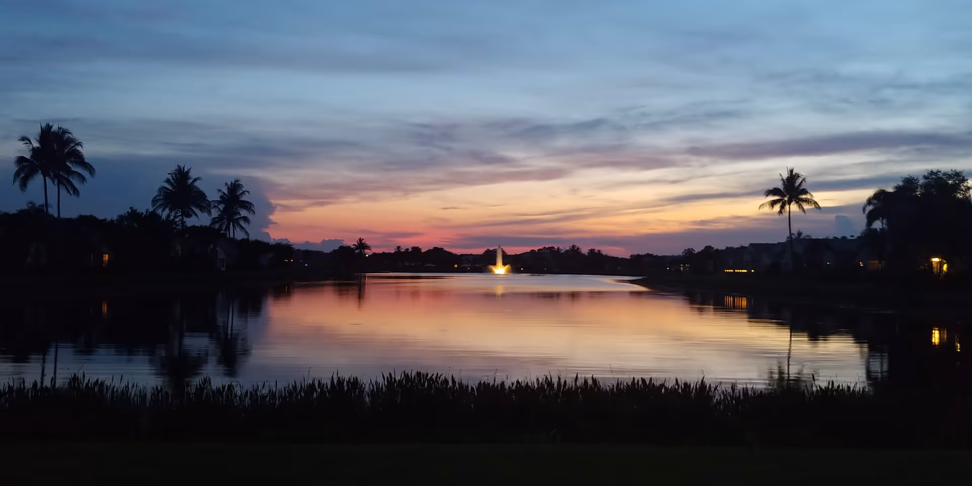 Sunset over a calm lake with silhouetted palm trees along the shore.