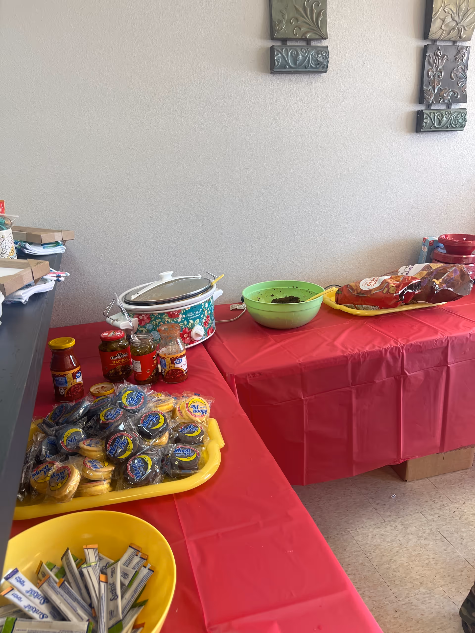 Long table with red cloth displays jars, condiments, and a slow cooker in a dining area