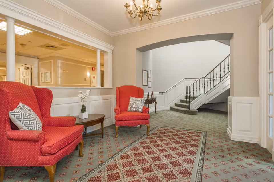 Elegant senior living lounge with red armchairs, chandelier, and carpeted seating area.