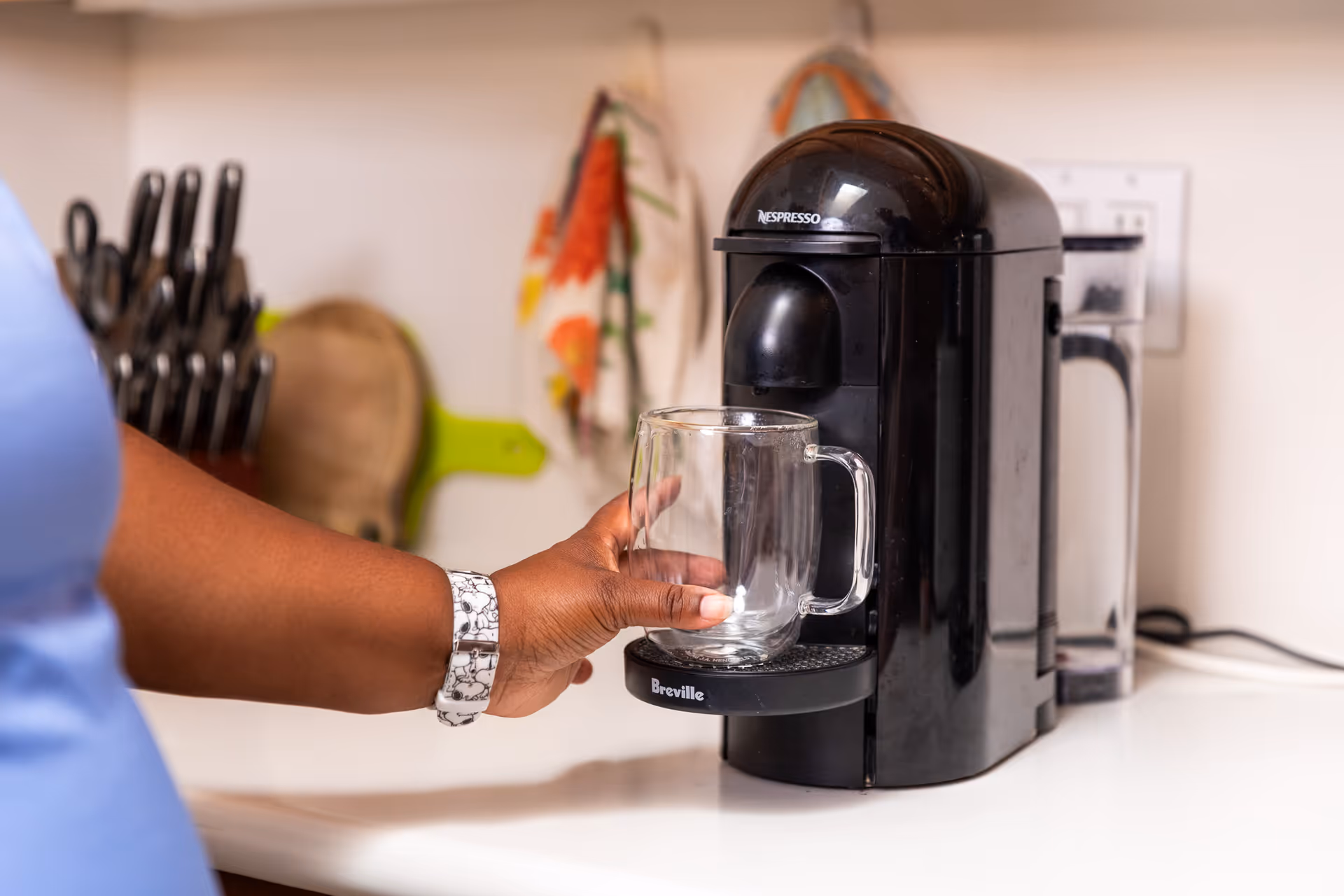 A person holds a glass mug under a black coffee maker on a kitchen counter.