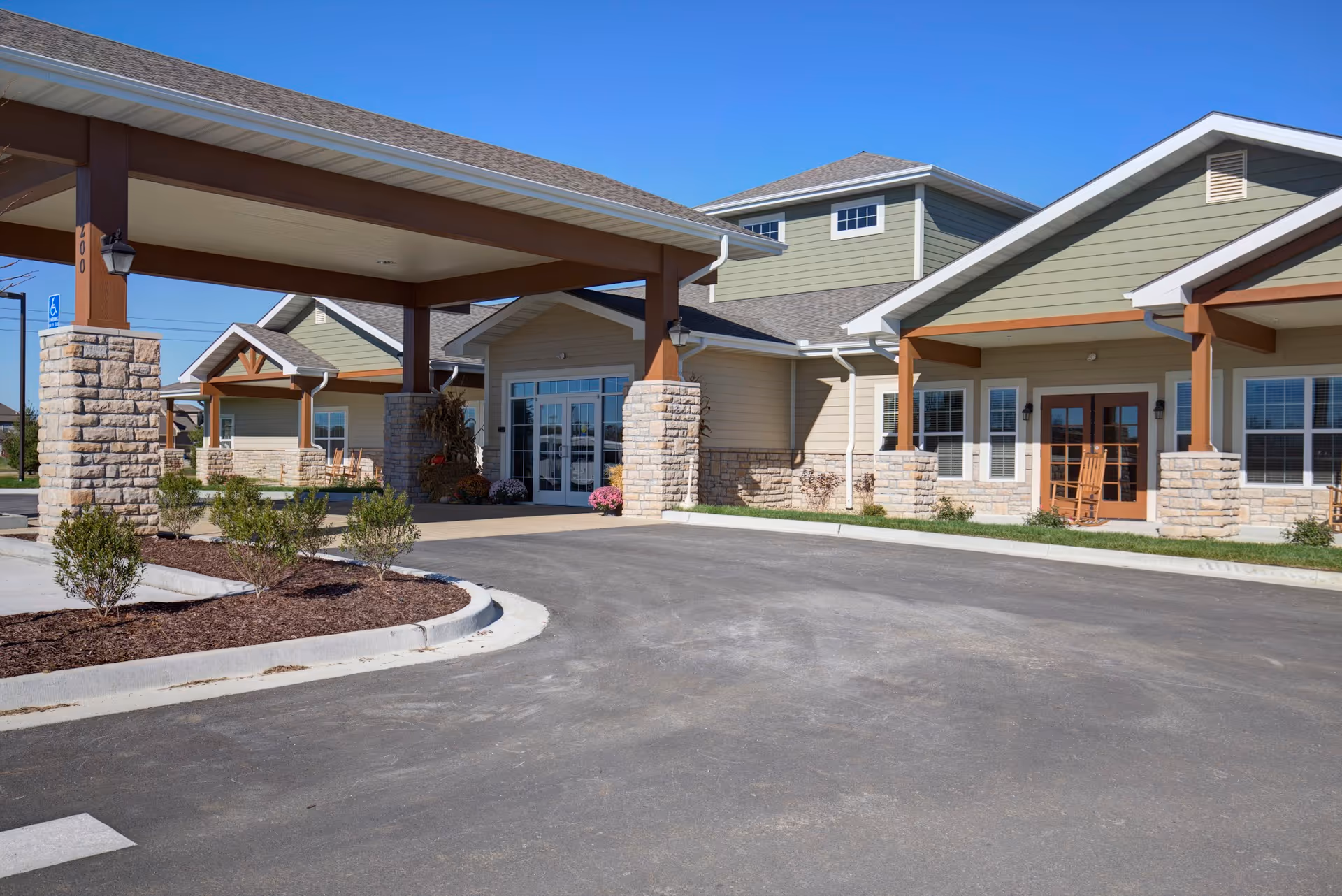 Exterior view of Oak Pointe of Kearney facility showing a covered entrance with stone pillars, green and beige siding, multiple windows, and a clear blue sky.