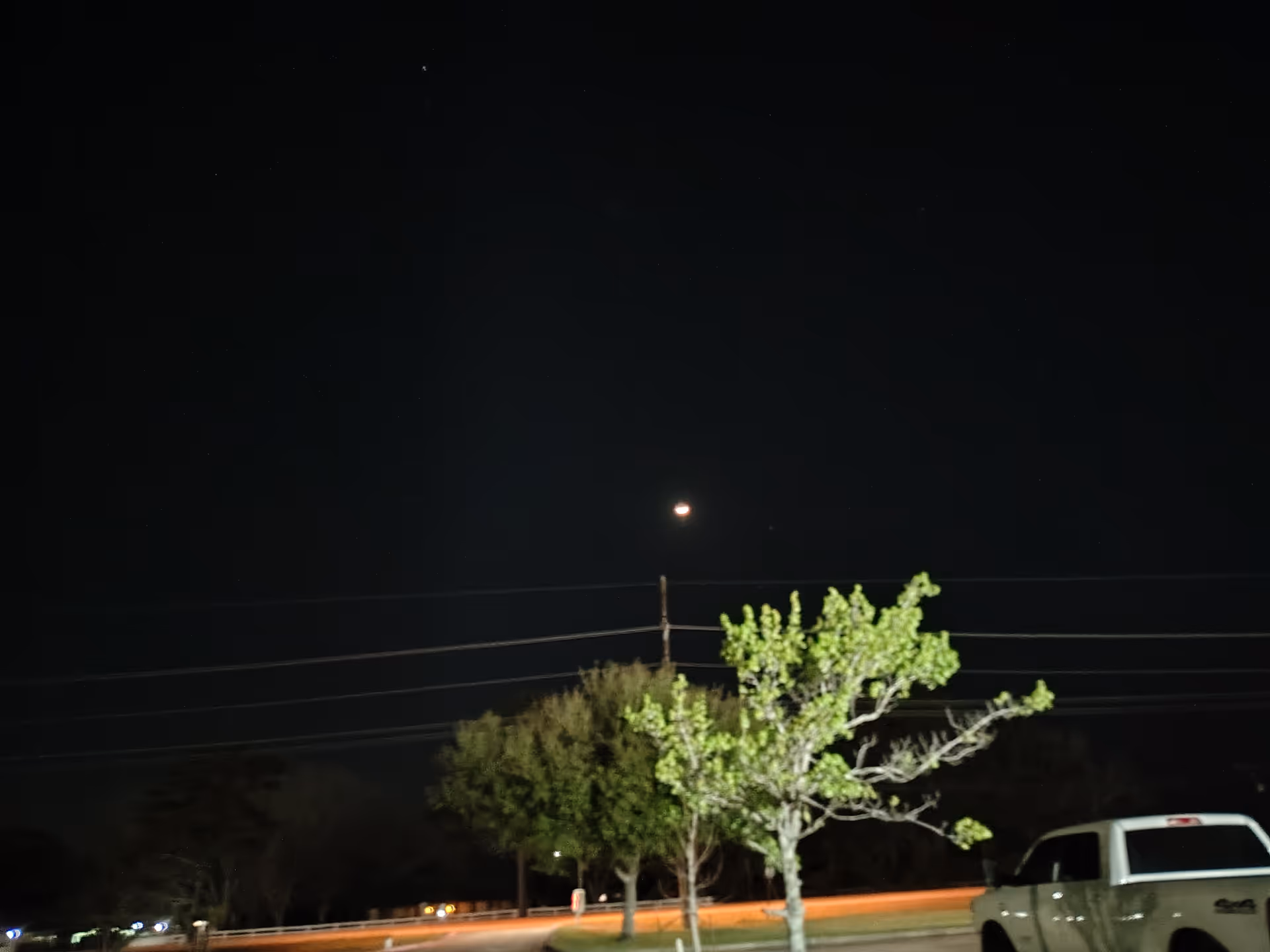 Night exterior view of a street with trees and a white pickup truck.