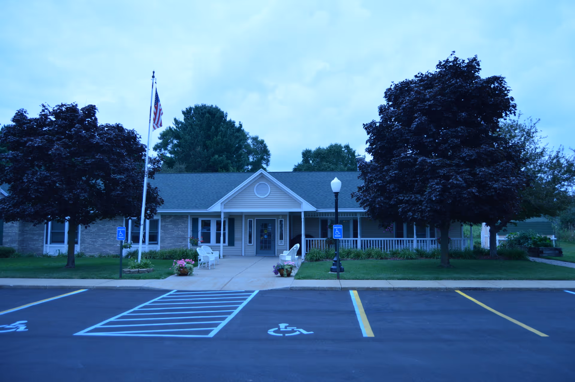 Front entrance of a single-story retirement facility with a flagpole, accessible parking spaces, and trees.