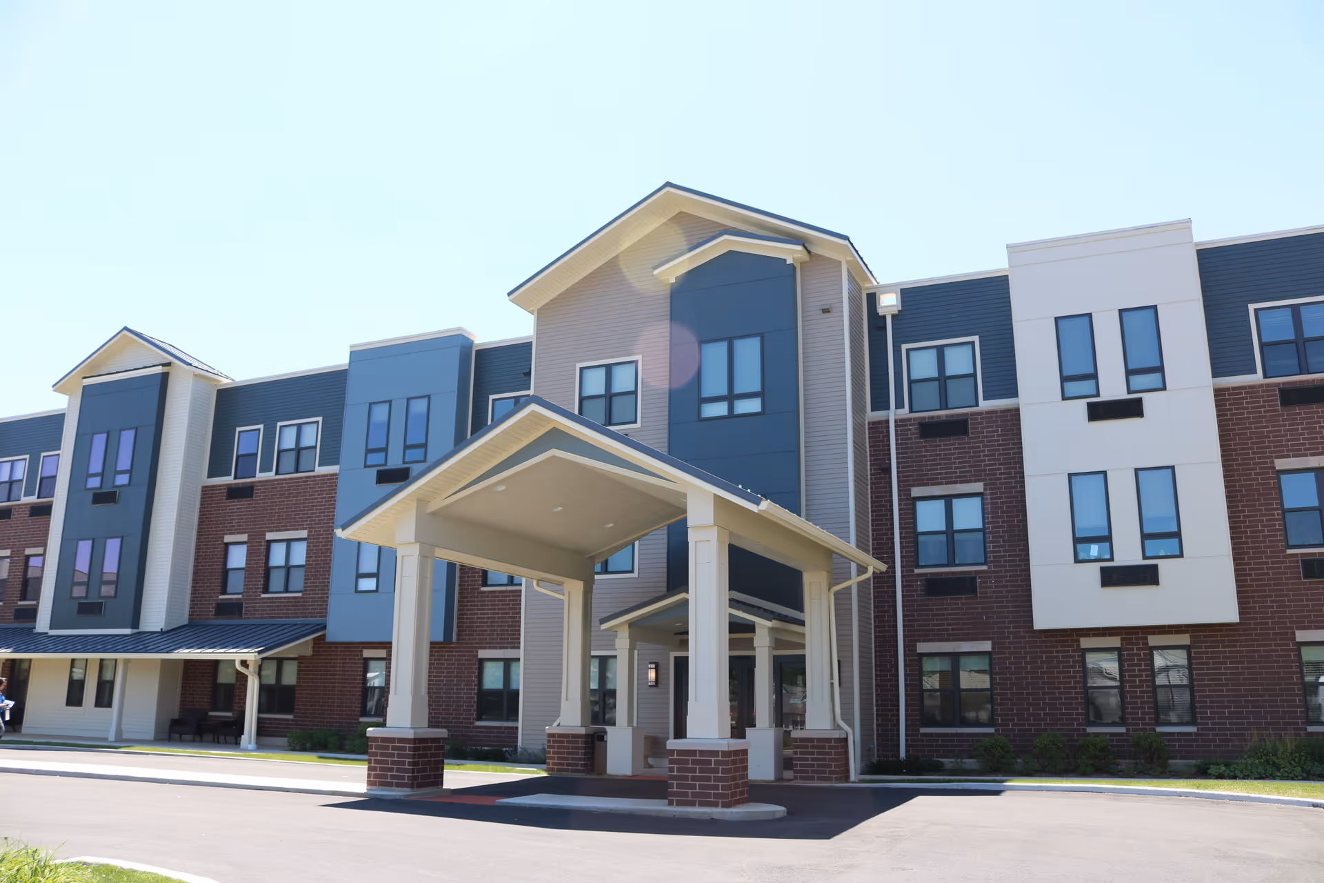 Front exterior view of a modern three-story senior living facility building with a covered entrance supported by columns. The building features a combination of brick, blue, and beige paneling with multiple windows under a clear blue sky.