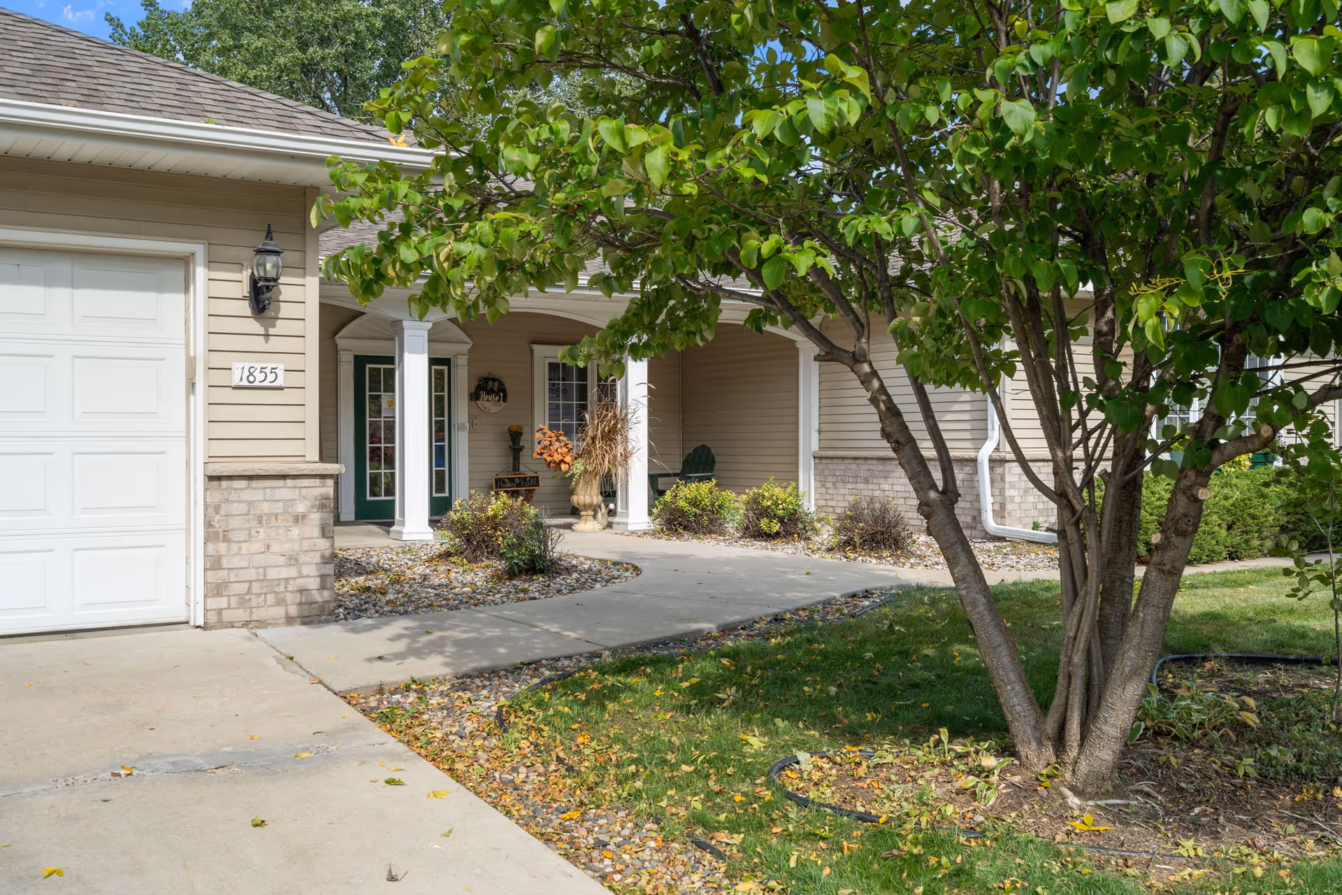 Front exterior view of a residential-style building with a garage on the left, a covered porch with white columns, and a tree with green leaves in the foreground. The building has beige siding and brick accents, with some landscaping including bushes and a small garden area.