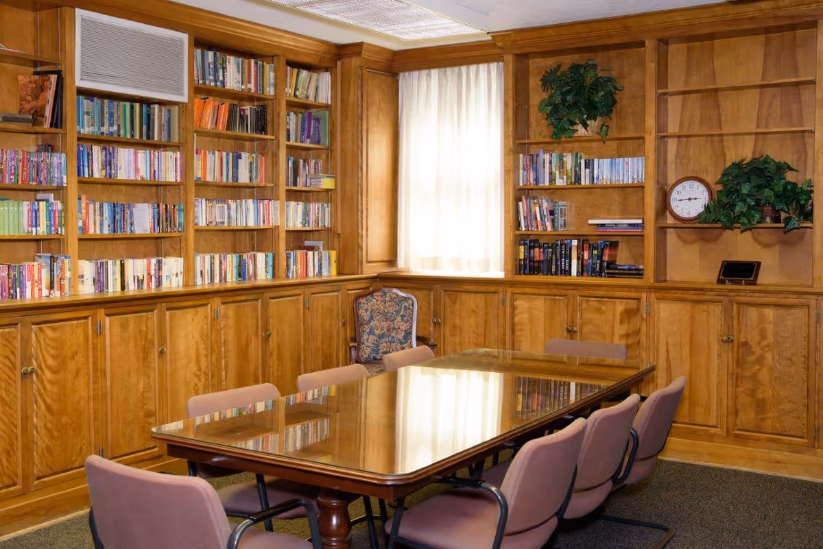 A cozy room with wooden bookshelves filled with books and some decorative plants. There is a large wooden table with a reflective surface surrounded by eight pink cushioned chairs. A window with sheer white curtains allows natural light into the room.