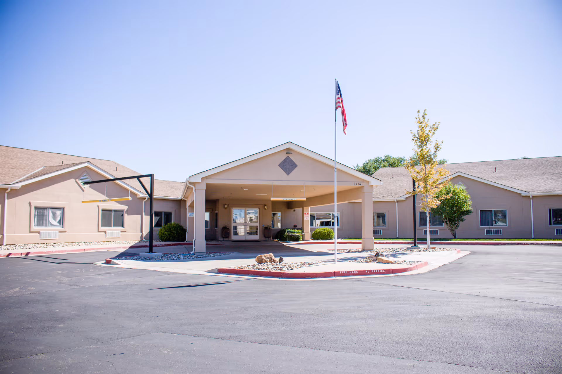 Exterior view of Vista Mesa Assisted Living facility showing a single-story building with a covered entrance, an American flag on a flagpole, and a paved driveway under a clear blue sky.