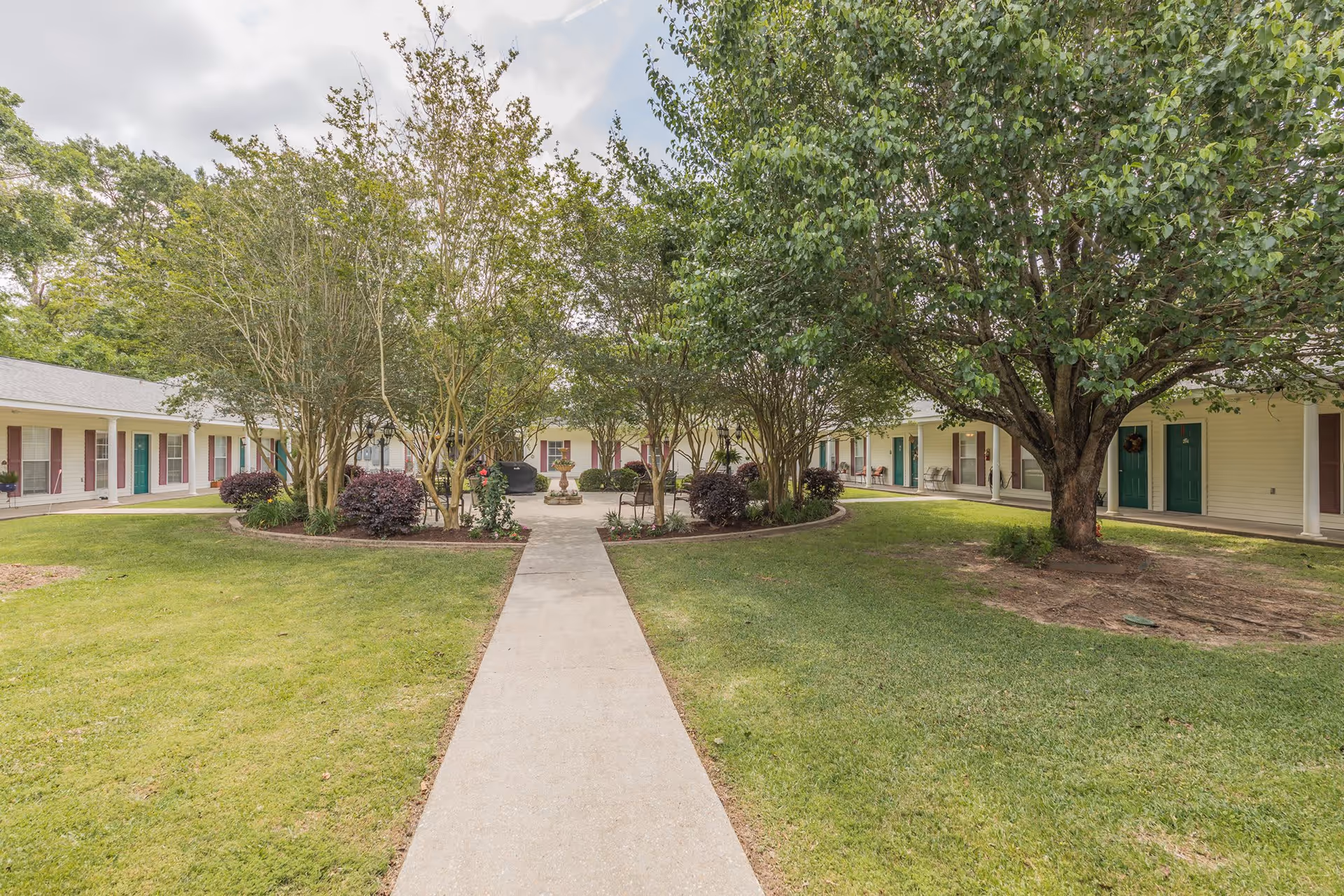 Concrete walkway through a grassy courtyard lined with trees leading to a single-story assisted living building with multiple doors and porches.