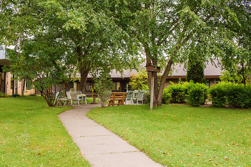 Path winds through a grassy courtyard with chairs and trees outside the facility.