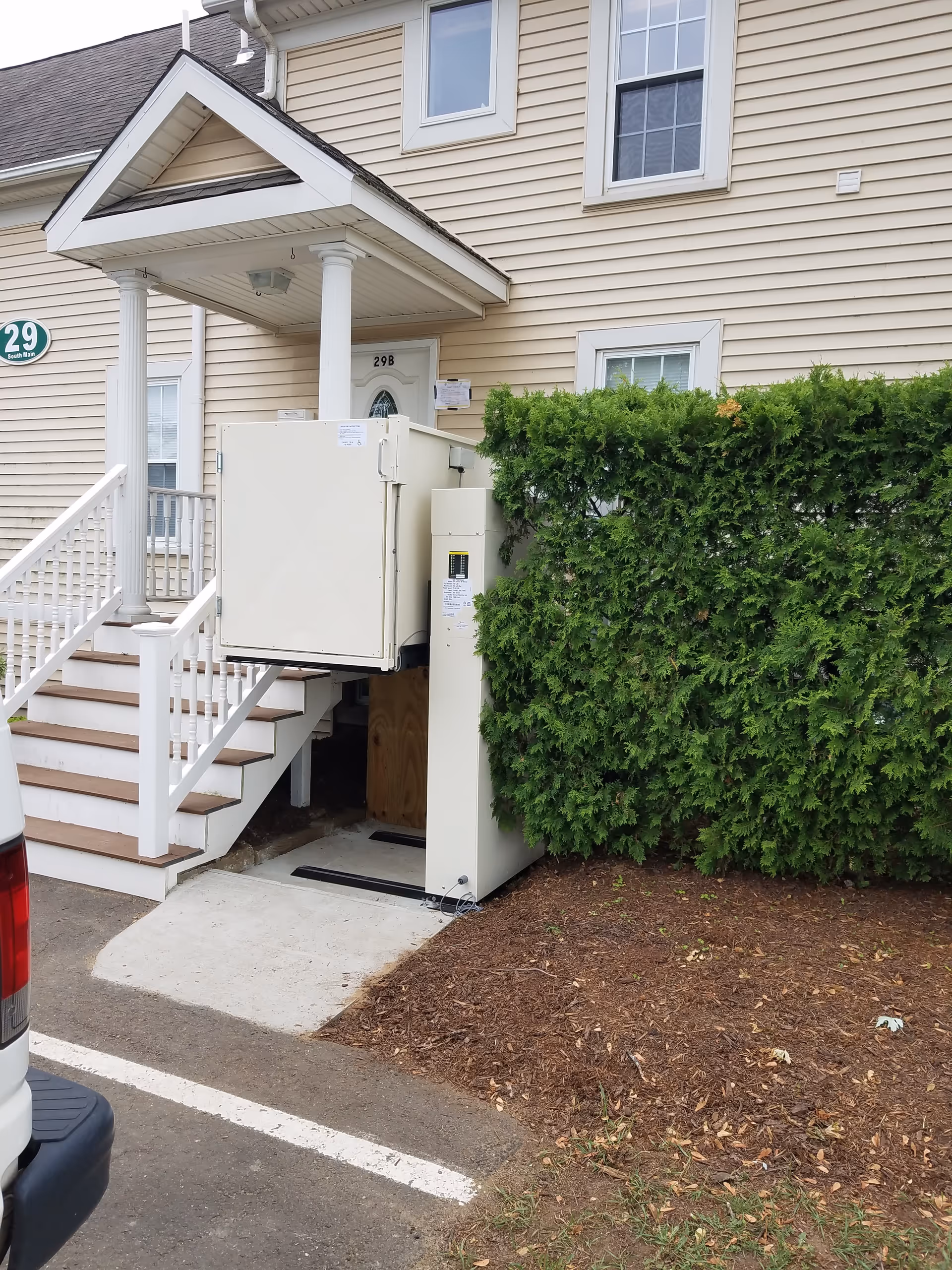 Exterior view of beige senior living building with stairs, railing, and a vertical lift beside hedges.