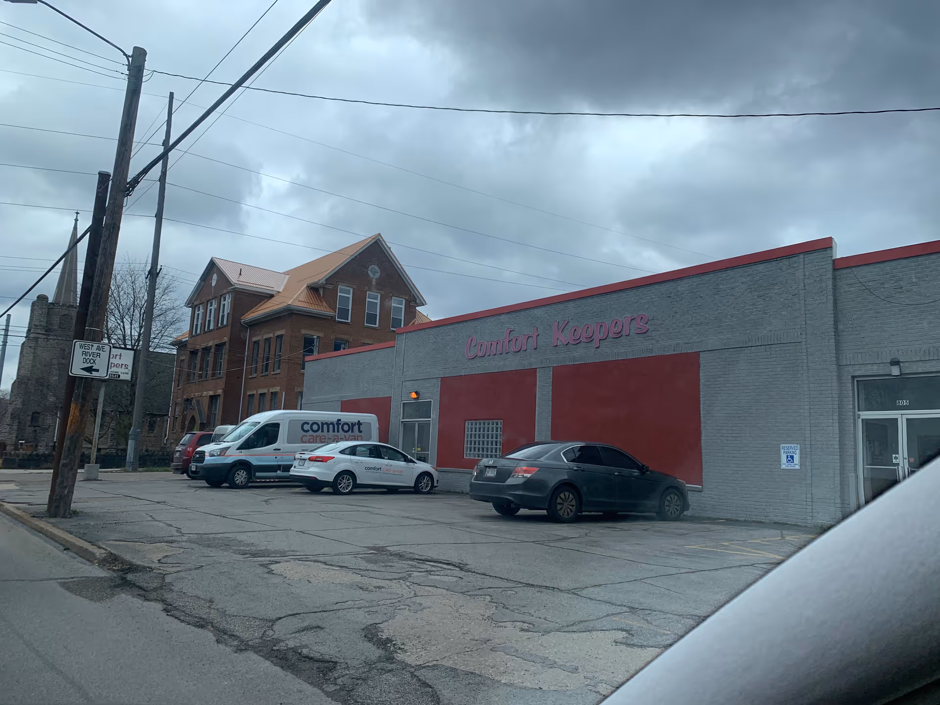Exterior view of Comfort Keepers facility with parked cars in front, cloudy sky.