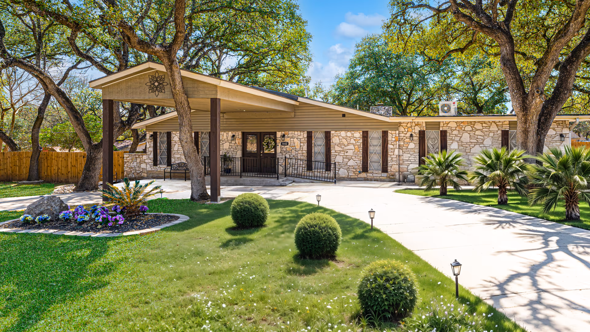 Stone-front single-story assisted living home with a covered entry, circular driveway, manicured lawn and trees.