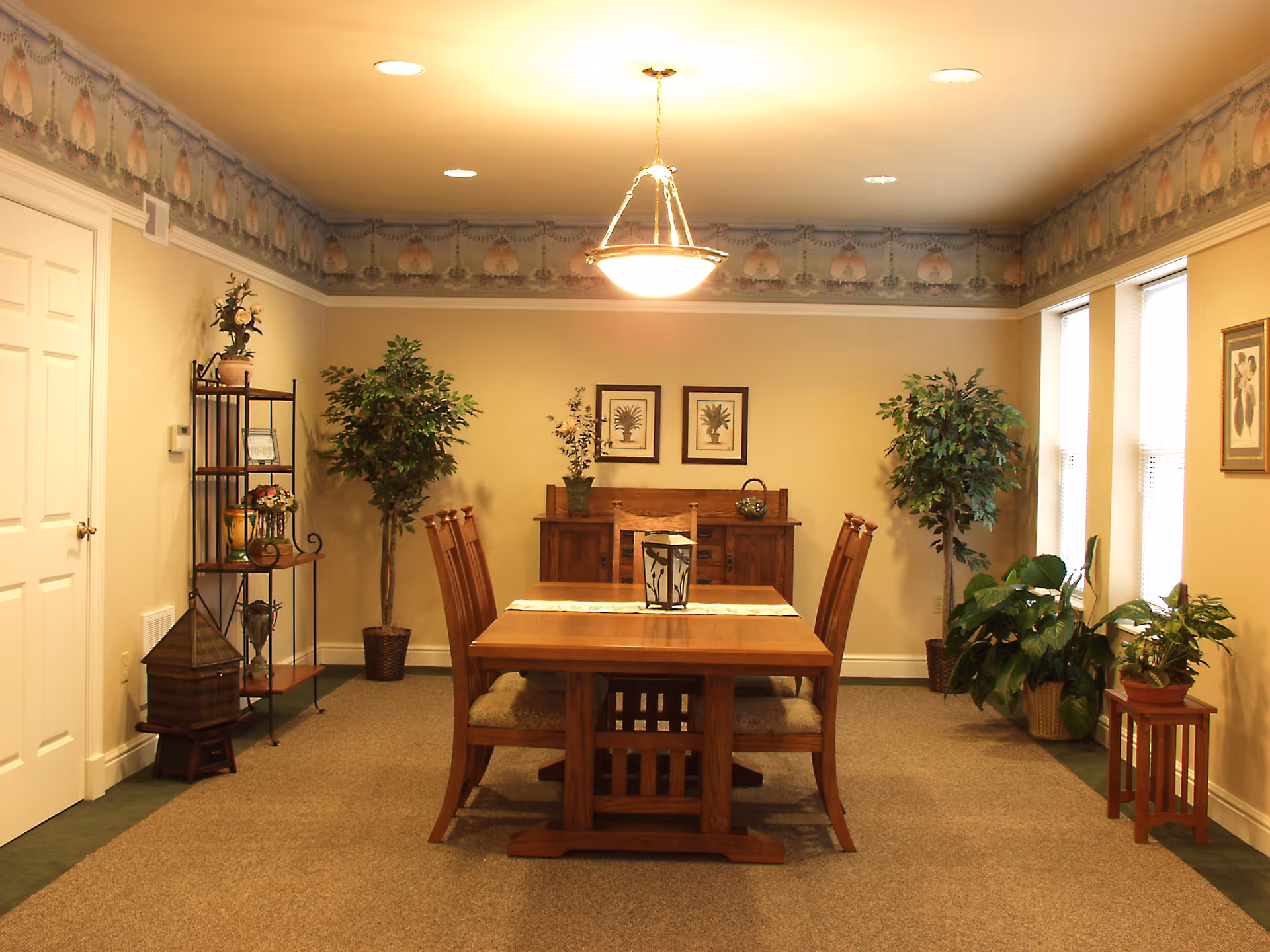 A warmly lit dining room with a wooden dining table and six chairs. The room features beige walls with a decorative border near the ceiling, two framed pictures on the back wall, and several potted plants placed around the room. A metal shelving unit with decorative items is on the left side, and three windows with blinds are on the right side.
