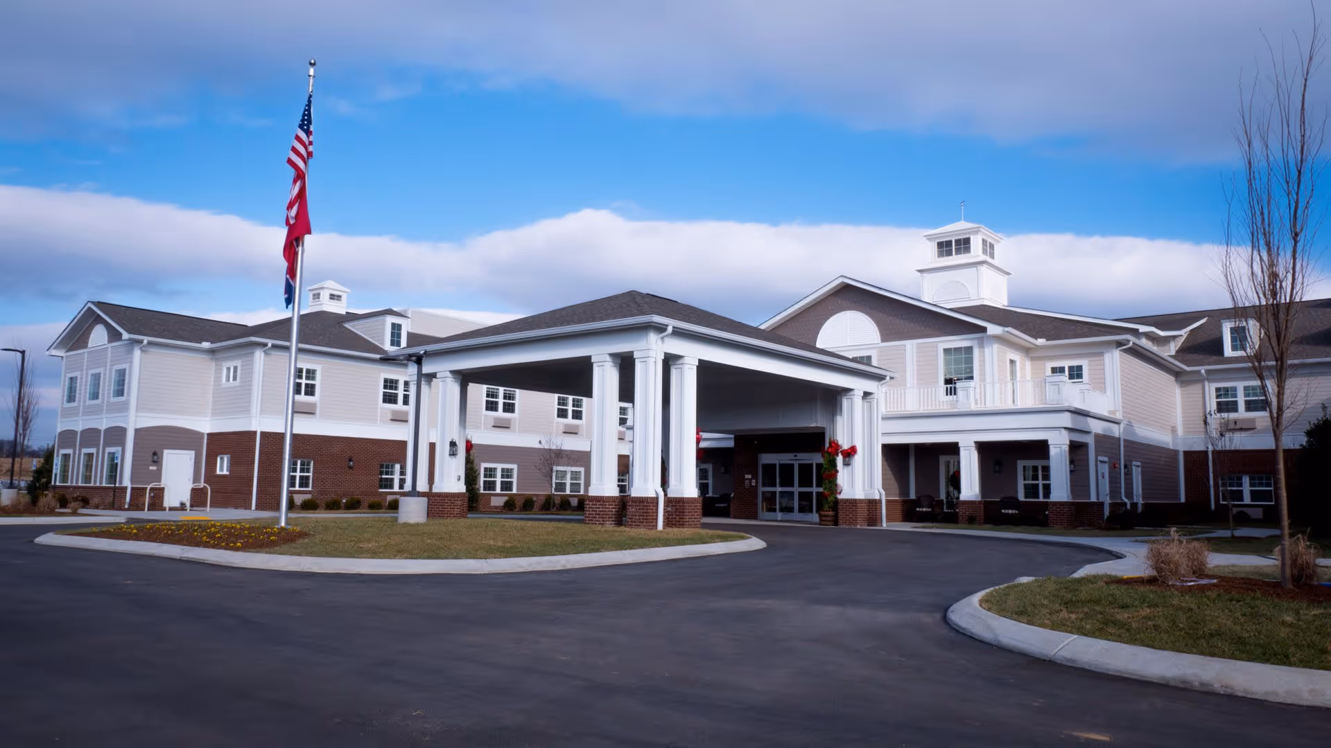 Exterior view of The Goldton at Spring Hill senior living facility showing a large two-story building with a covered entrance, multiple windows, and a flagpole with American and state flags. The sky is partly cloudy and the driveway curves around landscaped areas with grass and small trees.