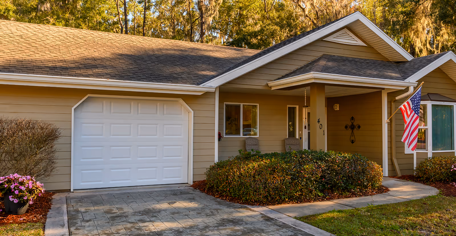 Exterior front of a single-story home with a garage door, covered porch, American flag and landscaped shrubs.