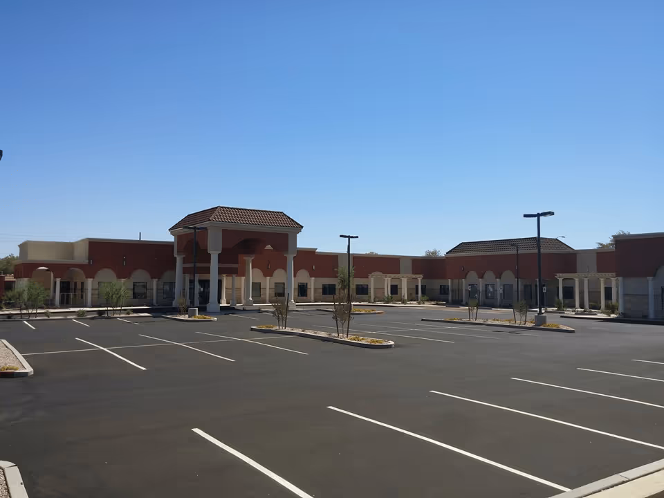 Empty parking lot in front of a single-story stucco building with a covered portico entrance under a clear blue sky.