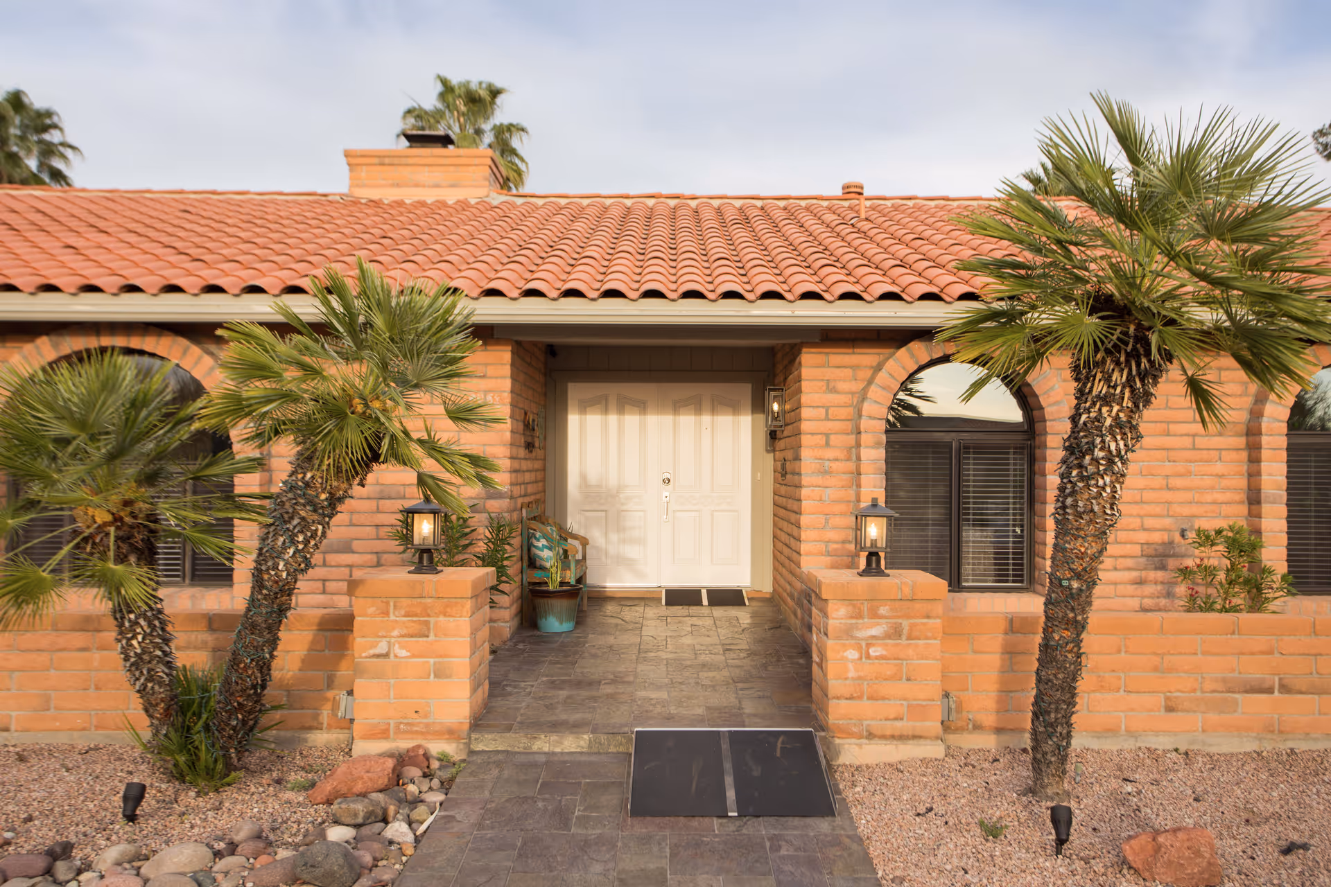 Front entrance of a brick building with a red tile roof, featuring a white double door, two small palm trees on either side, stone pathway with a ramp, and two lantern-style lights on brick pillars.
