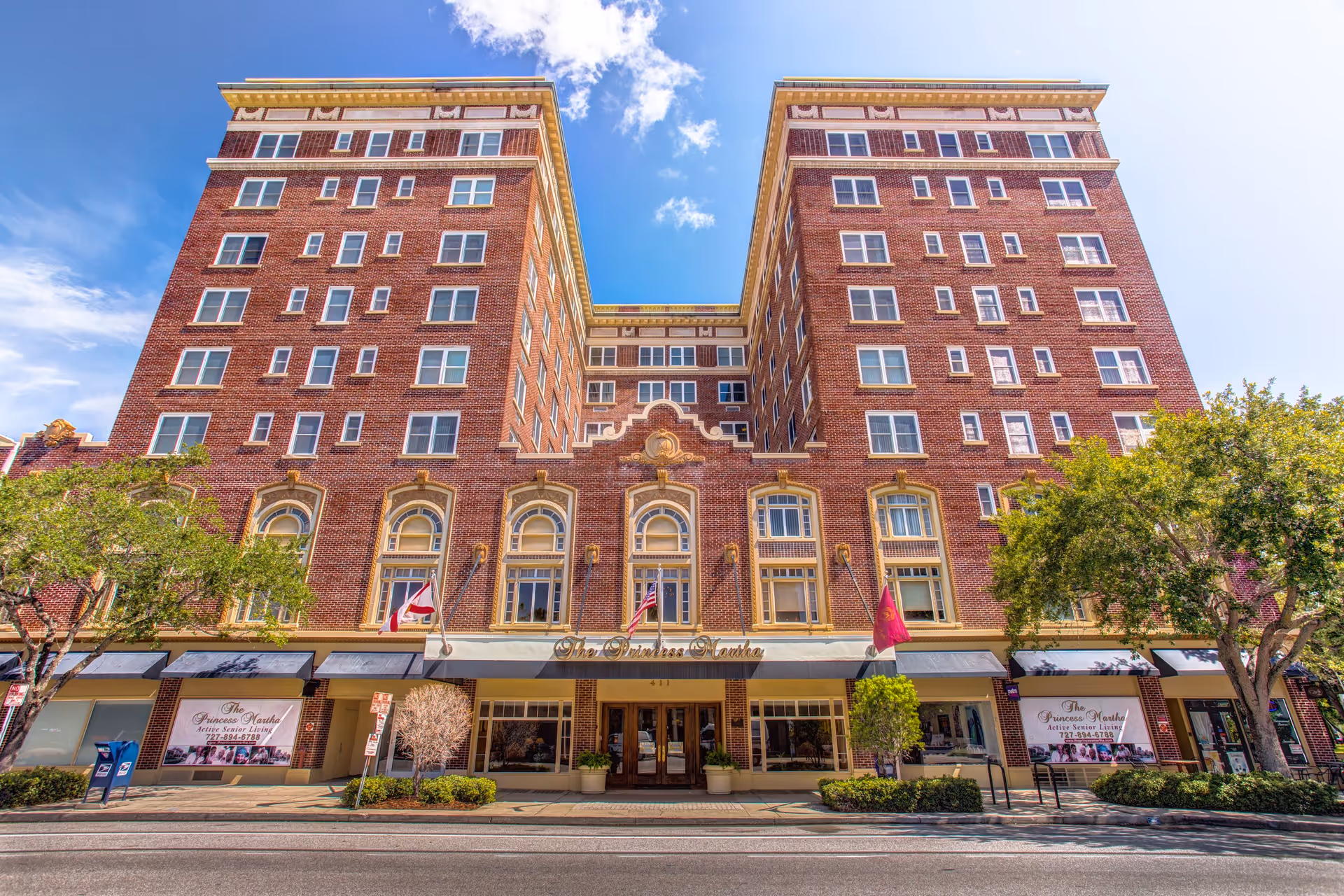 Front façade of The Princess Martha, a multi-story red brick building with ground-level awnings, flags, and trees along the sidewalk.