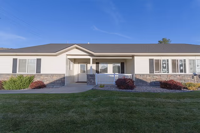 Single-story beige memory care building with a covered central entrance, small porch railing, stone accents and a manicured lawn under a clear blue sky.